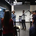 A student holds up a mic as his classmates record a voiceover in front of a TV