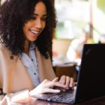 Black woman happily working on her computer in a cafe.