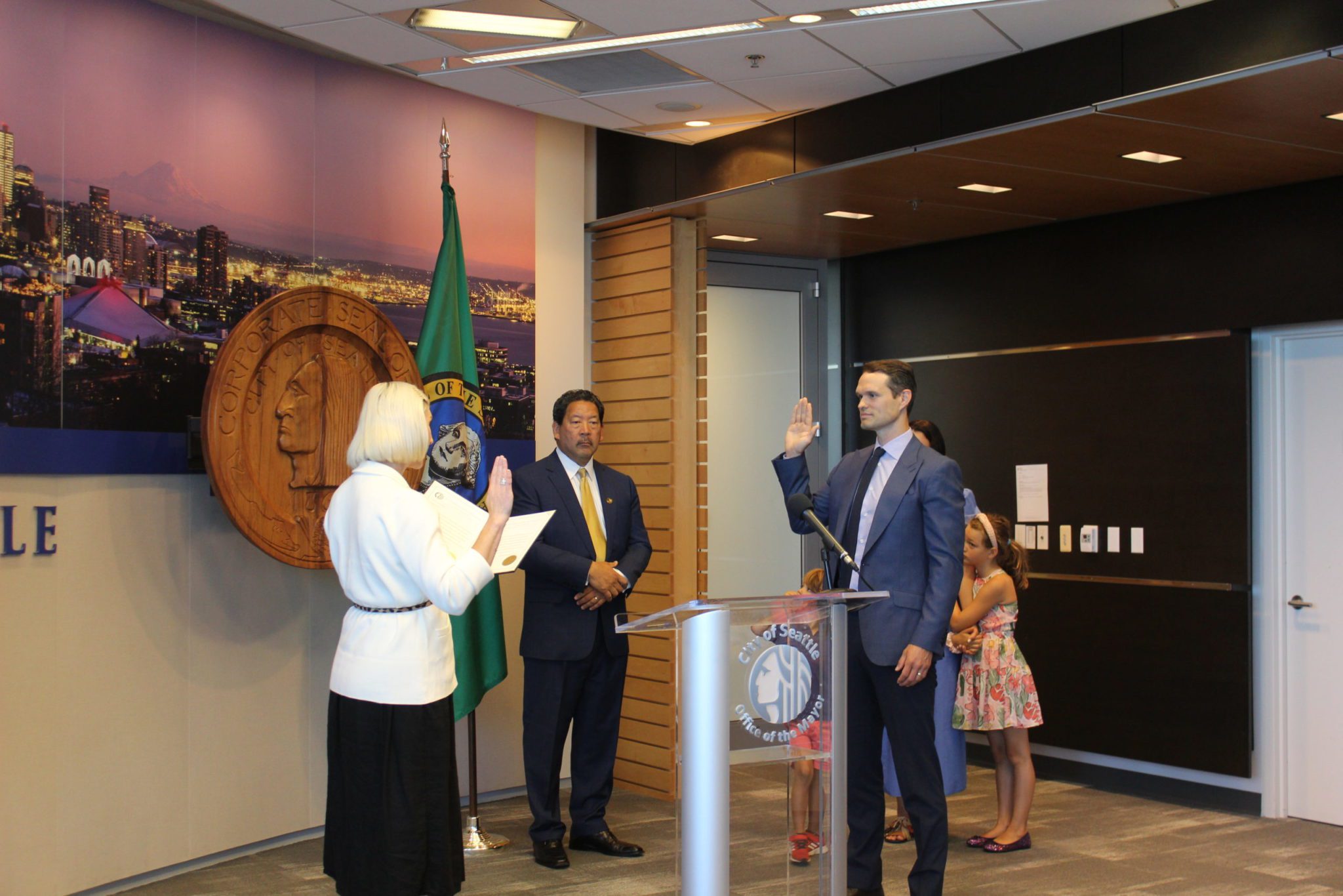 Former OED director Markham McIntyre being sworn into office. McIntyre is standing with his right hand raised, being sworn in by Seattle Council President, with former Mayor Bruce Harrell to his right.