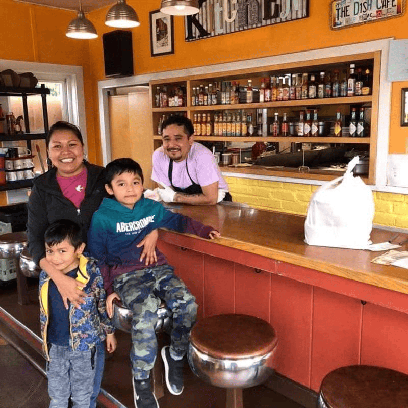 Two individuals and two children standing inside a restaurant near the counter, with shelves of condiments in the background.