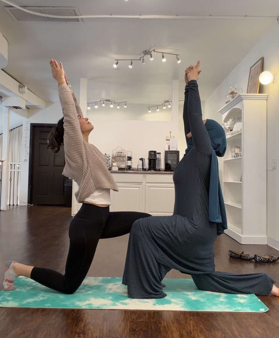 Two individuals on a yoga mat indoors, kneeling in a mirrored pose with arms raised overhead.