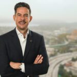 OED Director Beto Yarce standing in front of a window smiling with his arms crossed. Behind him is southern view of the City of Seattle. He is wearing a dark suit and white open collared shirt.