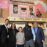 Five people pose for a photo at Le's Deli and Bakery in Seattle's Othello neighborhood. They are standing in front of a mural and a shelf of home baked rice crispy treats. From left to right the people are: OED Director Beto Yarce, Mayor Katie Wilson, business owner Cindy Ly, RVCDF director Dr. Alvertis Brooks Jr., and Senior Director at Grow America Chuck Depew.