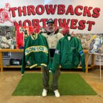 Man holds vintage SuperSonics and Seahawks satin jackets in front of a "Throwbacks Northwest" banner and paintings of Seattle athletes.