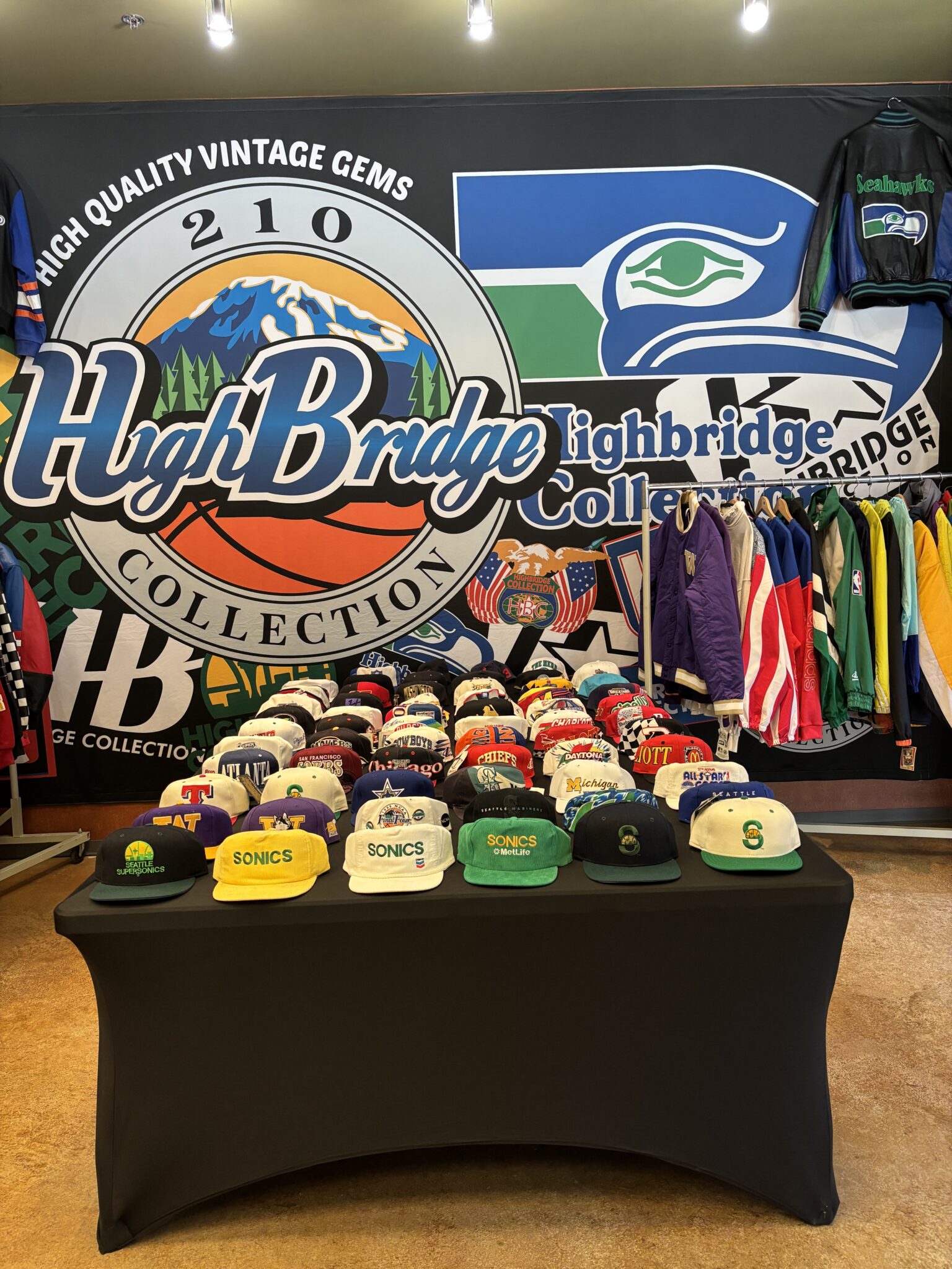 Vintage sports apparel booth with a "High Bridge Collection" backdrop, a rack of colorful jackets, and a table of team snapback hats.